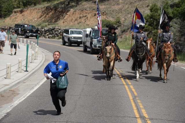An Explorer jogs ahead of the mounted unit carrying the United States and Colorado flags on a mountain parade route, sheriff vehicles following