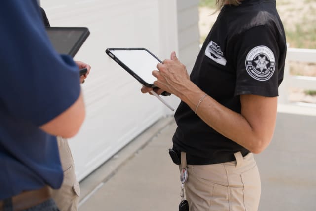 Close-up of an assessor's hands recording notes on a tablet with a stylus outside a garage door, a colleague's tablet in the foreground