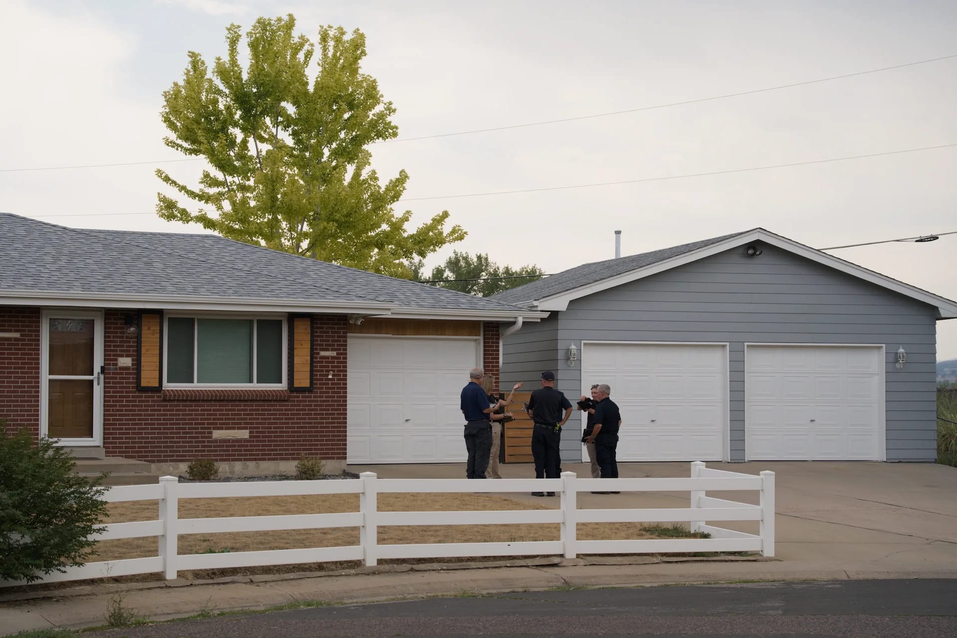 Five assessors talk in the driveway of a brick ranch home with a white picket fence, a tall tree and garage behind them
