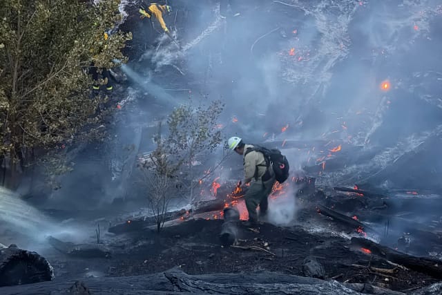 A wildland firefighter in a white helmet and pack works through smoke and glowing embers among charred logs, a second firefighter working upslope