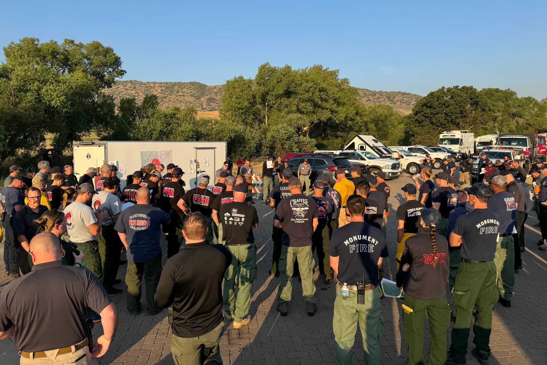 Dozens of firefighters from multiple agencies stand in a circle around a map on a trailer for a morning briefing, trucks and foothills behind