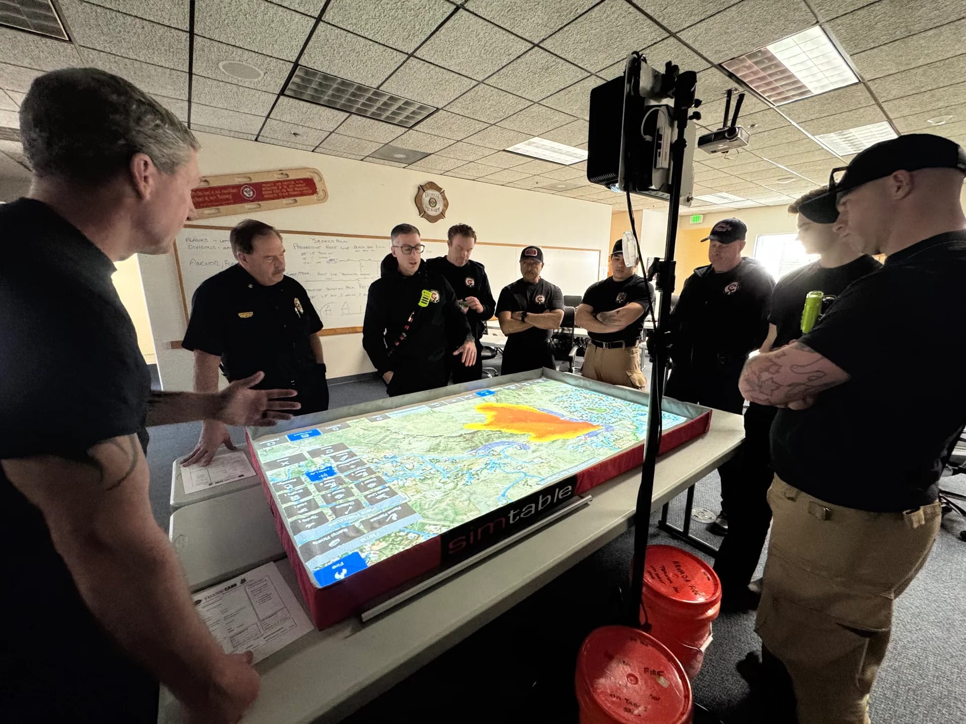 Arvada Fire firefighters gather around a SimTable, a projected terrain map showing a simulated wildfire spreading in orange, while an instructor gestures toward the scenario