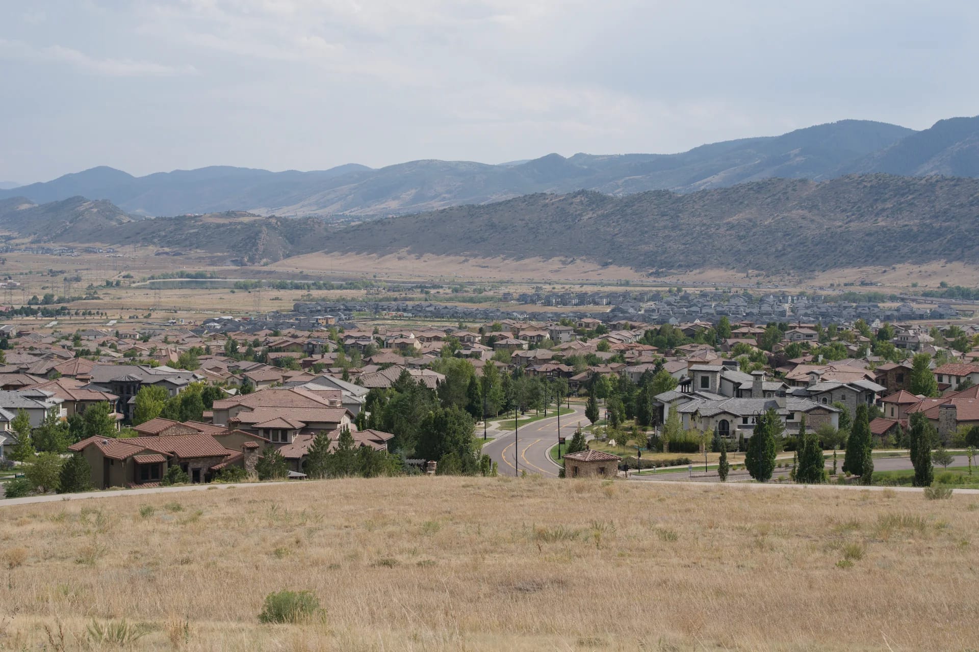 A foothills neighborhood of tile-roofed homes seen from a dry grassy hilltop, with a hogback ridge and mountains behind