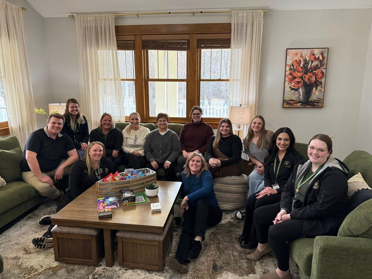 A group of eleven staff members sit together in a bright, cozy living room with green couches and large windows. They are smiling toward the camera, with board games and snacks on a wooden coffee table in front of them.
