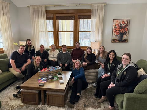 A group of eleven staff members sit together in a bright, cozy living room with green couches and large windows. They are smiling toward the camera, with board games and snacks on a wooden coffee table in front of them.
