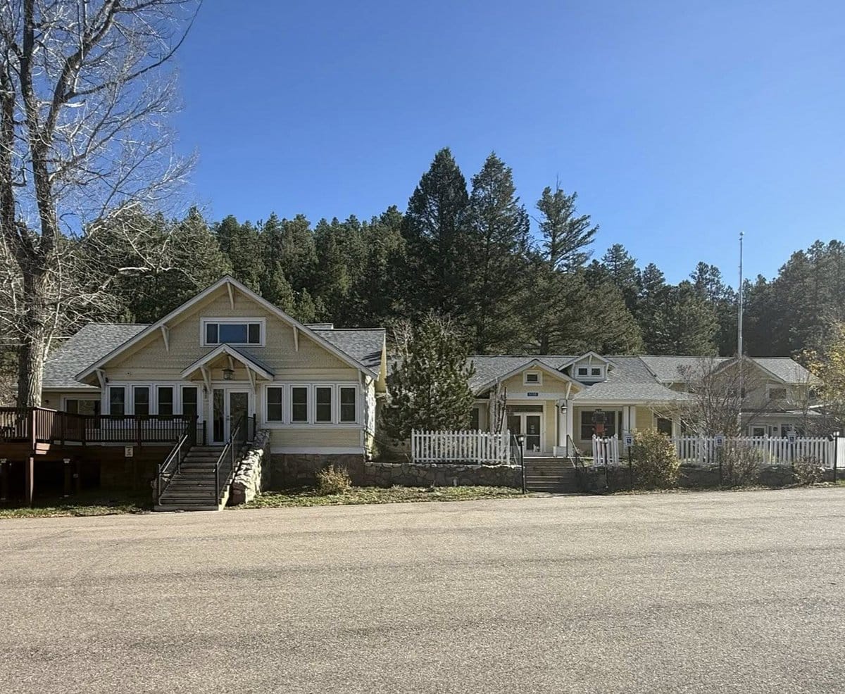 A light-yellow house with white trim sits at the base of a wooded hillside under a clear blue sky. A white picket fence lines the front entrance, and tall pine trees rise behind the building.