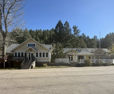 A light-yellow house with white trim sits at the base of a wooded hillside under a clear blue sky. A white picket fence lines the front entrance, and tall pine trees rise behind the building.