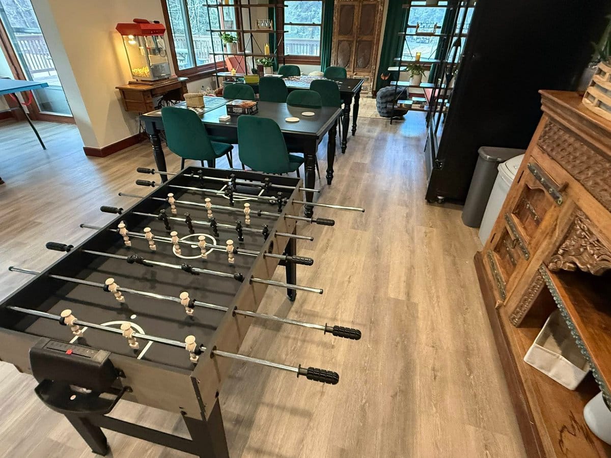 A recreation room with a foosball table in the foreground and two tables with green chairs behind it. A popcorn machine sits on a small wooden cabinet near large windows overlooking trees.