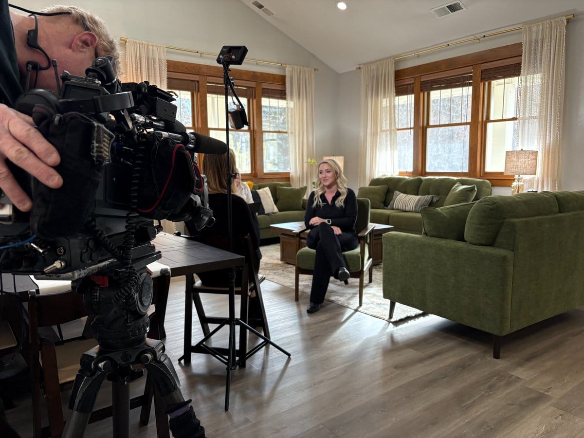 A woman sits in a green chair in a bright living room speaking to a tv reporter. A cameraman with a video camera and lighting equipment are set up in the foreground, filming her as she speaks.