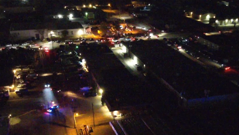 Nighttime drone view of a large car meetup filling a commercial parking lot, with law enforcement vehicles staged nearby