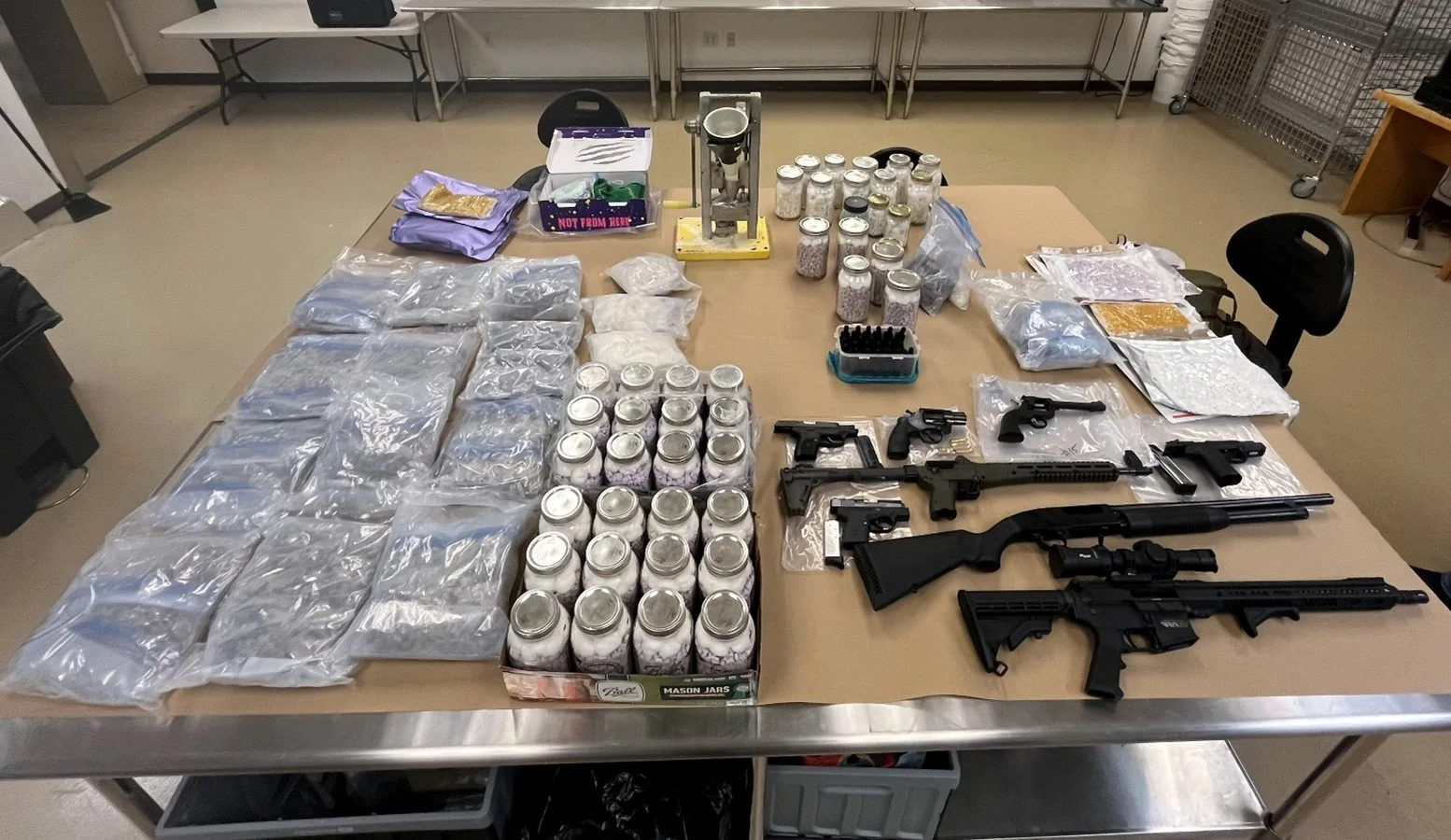 Evidence table from the Covelli investigation covered with bagged narcotics, jars of pills and mushrooms, a pill press, and eight seized firearms