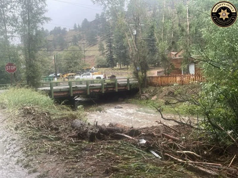 Flood debris piled along Bear Creek at a small bridge in Kittredge, with road crews and heavy equipment staged behind it