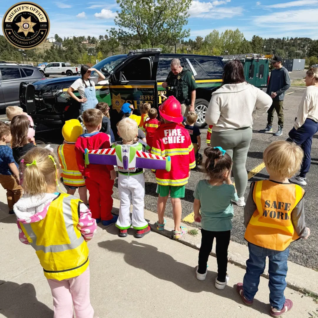 Deputy Bybee beside the open door of his patrol SUV as preschoolers in costumes and safety vests, including a fire chief and a space ranger, gather around him at Kids Garden