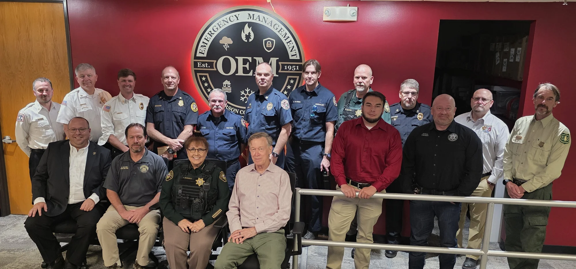 U.S. Senator John Hickenlooper in a group photo with JCSO, Emergency Management and local fire leaders in front of a red wall bearing the Emergency Management OEM seal