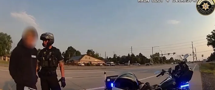 Body camera still of a JCSO motor deputy standing with the motorcycle rider, whose face is blurred, beside the road with the deputy's patrol motorcycle and its blue lights in the foreground