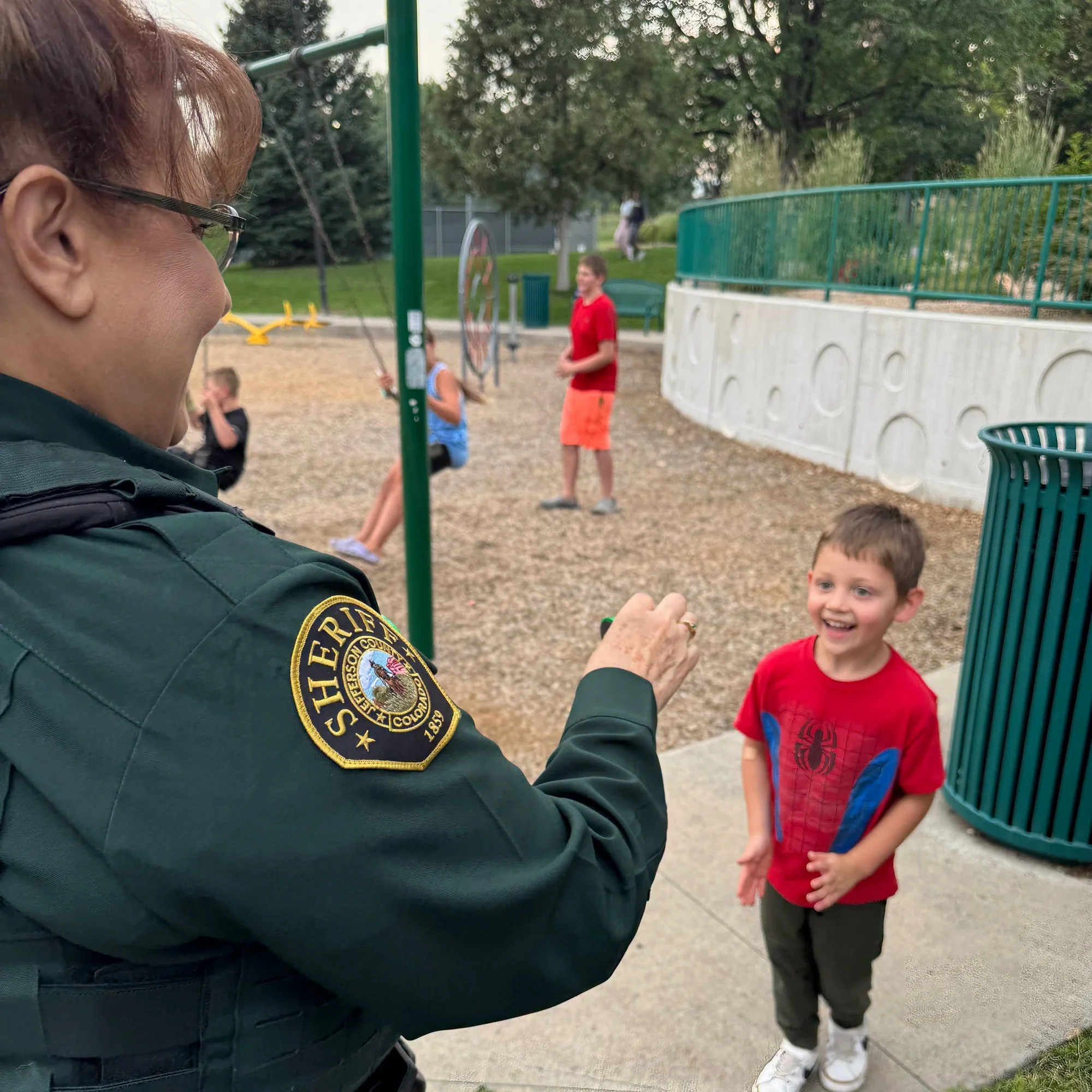 A JCSO deputy, seen over her shoulder with the Sheriff patch on her sleeve, greets a laughing young boy in a red Spider-Man shirt at a neighborhood playground