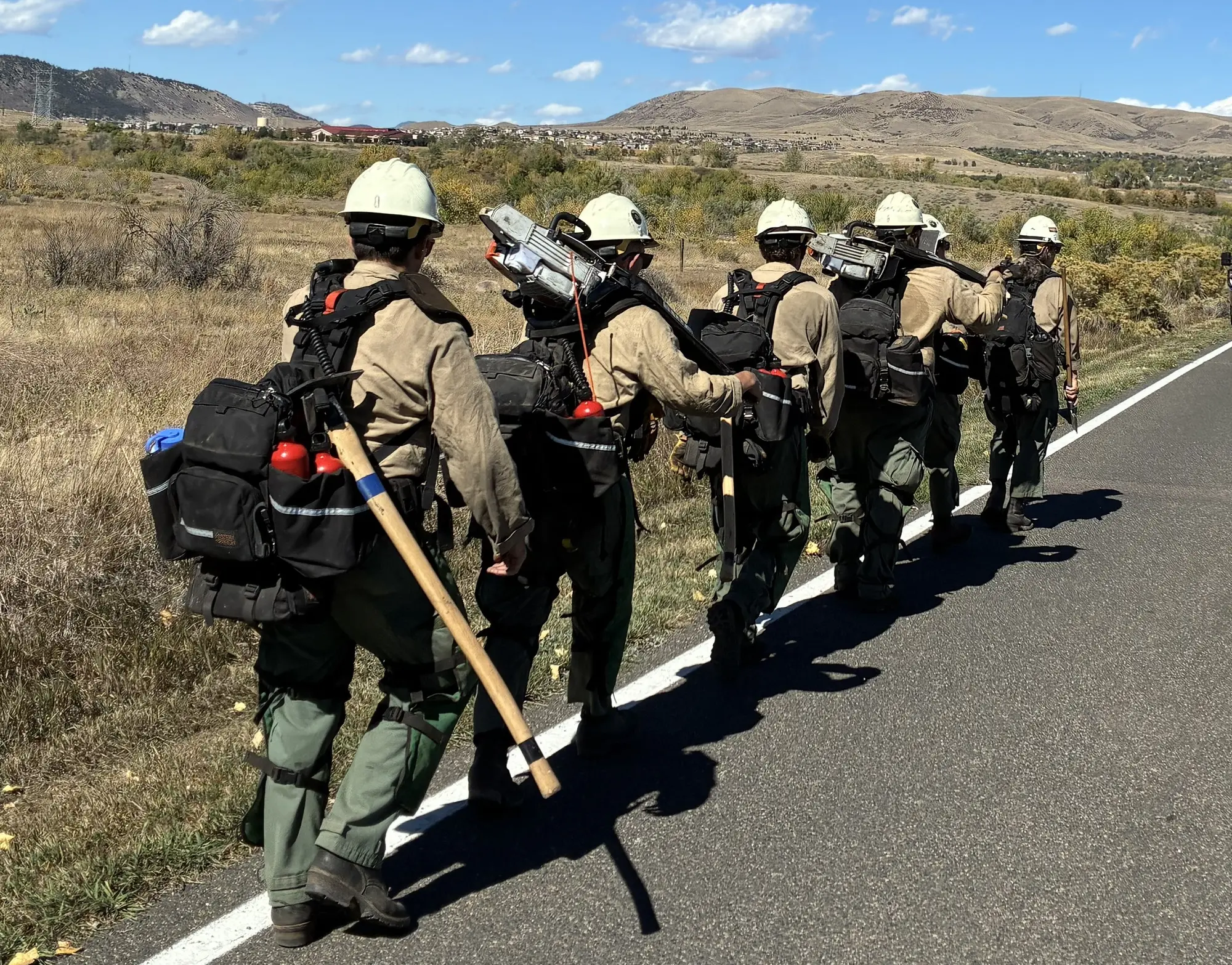 A wildland fire crew in white helmets and packs, carrying chainsaws and hand tools, walking single file along a road shoulder through dry foothills grassland