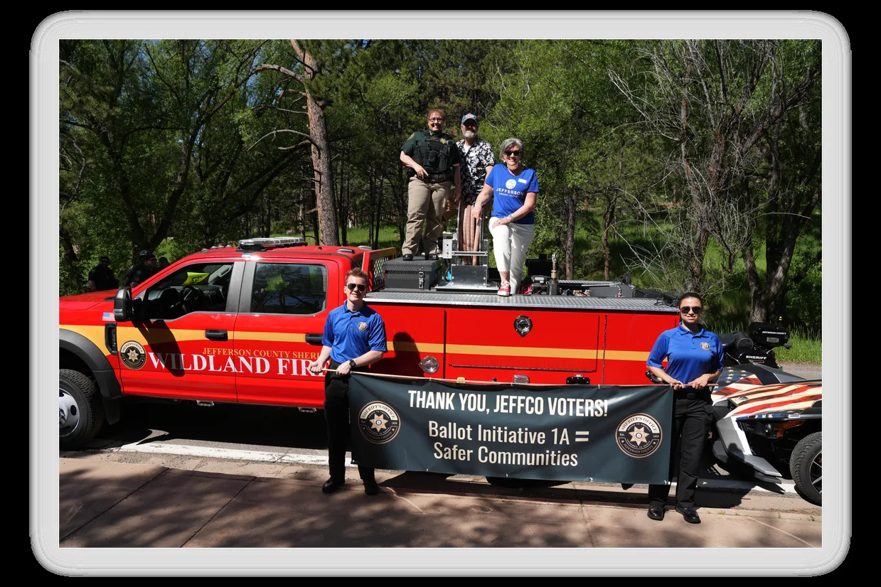 Two JCSO Explorers hold a banner reading Thank you, Jeffco voters! Ballot Initiative 1A = Safer Communities in front of a red Jefferson County Sheriff wildland fire brush truck; Sheriff Marinelli and two County Commissioners stand on the truck bed at the Evergreen Rodeo Parade.